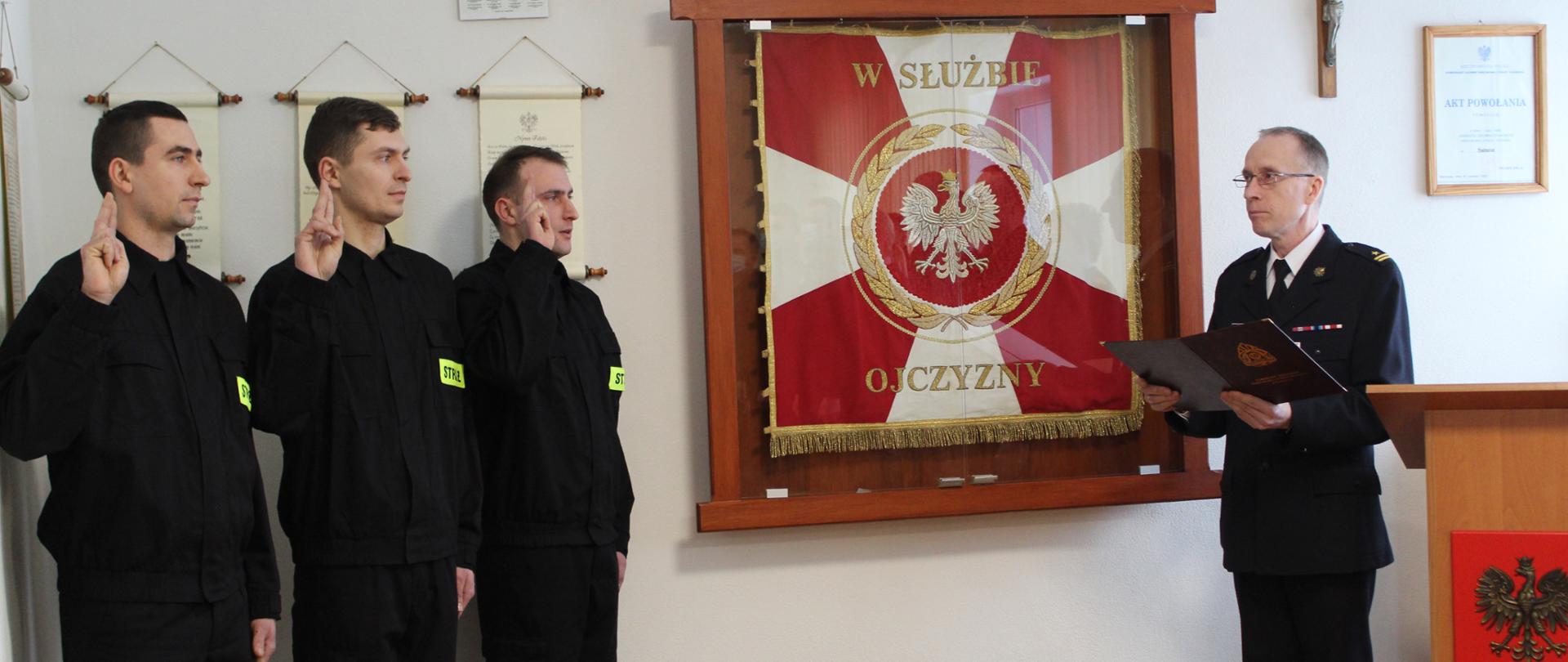 Z lewej strony zdjęcia widzimy trzech młodych mężczyzn z uniesionymi prawymi rękami do przysięgi ubranych w czarne ubrania koszarowe z jasnożółtymi emblematami, z czarnym napisem "STRAŻ". Po lewej stronie stoi dojrzały mężczyzna ubrany w białą koszulę z czarnym krawatem, marynarką czarną, trzymający w ręku dokumenty. Po jego prawej stronie stoi mównicza z godłem Rzeczypospolitej Polskiej. W tle widzimy Sztandar KP PSP Świdwin, Krzyż. 