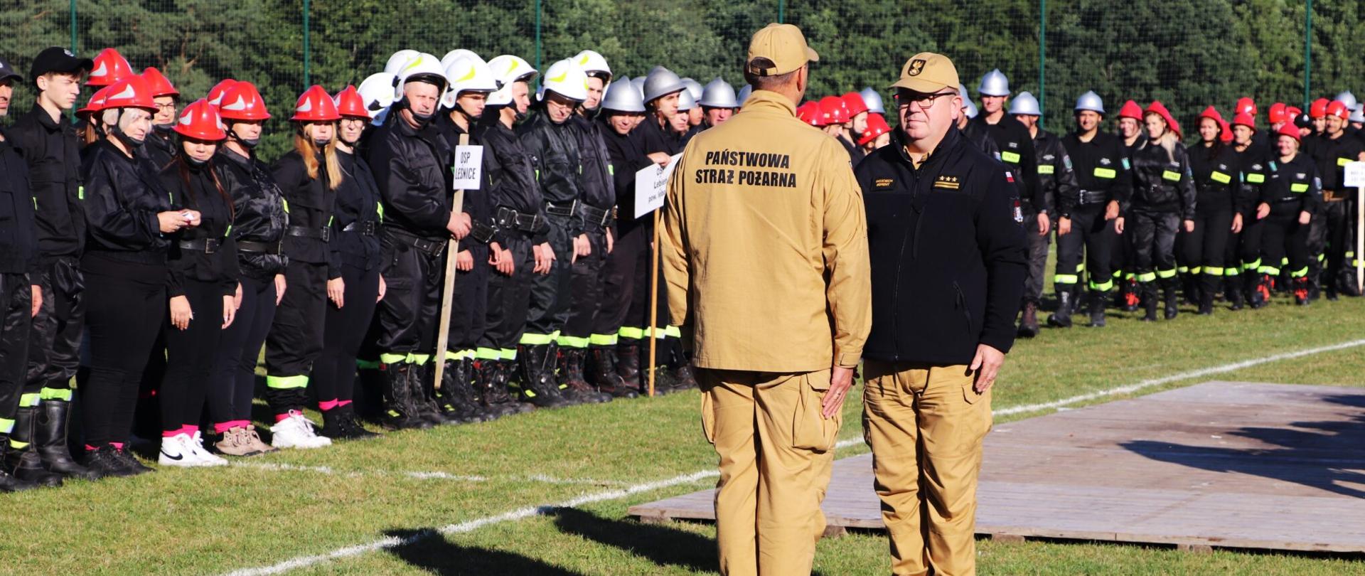 Zastępca Pomorskiego Komendanta Wojewódzkiego Państwowej Straży Pożarnej starszy brygadier Wojciech Berent przyjmuje meldunek od koordynatora zawodów starszego kapitana Marcina Elwart. W tle widoczne drużyny Ochotniczych Straży Pożarnych stojące w dwóch rzędach na boisku sportowym. 