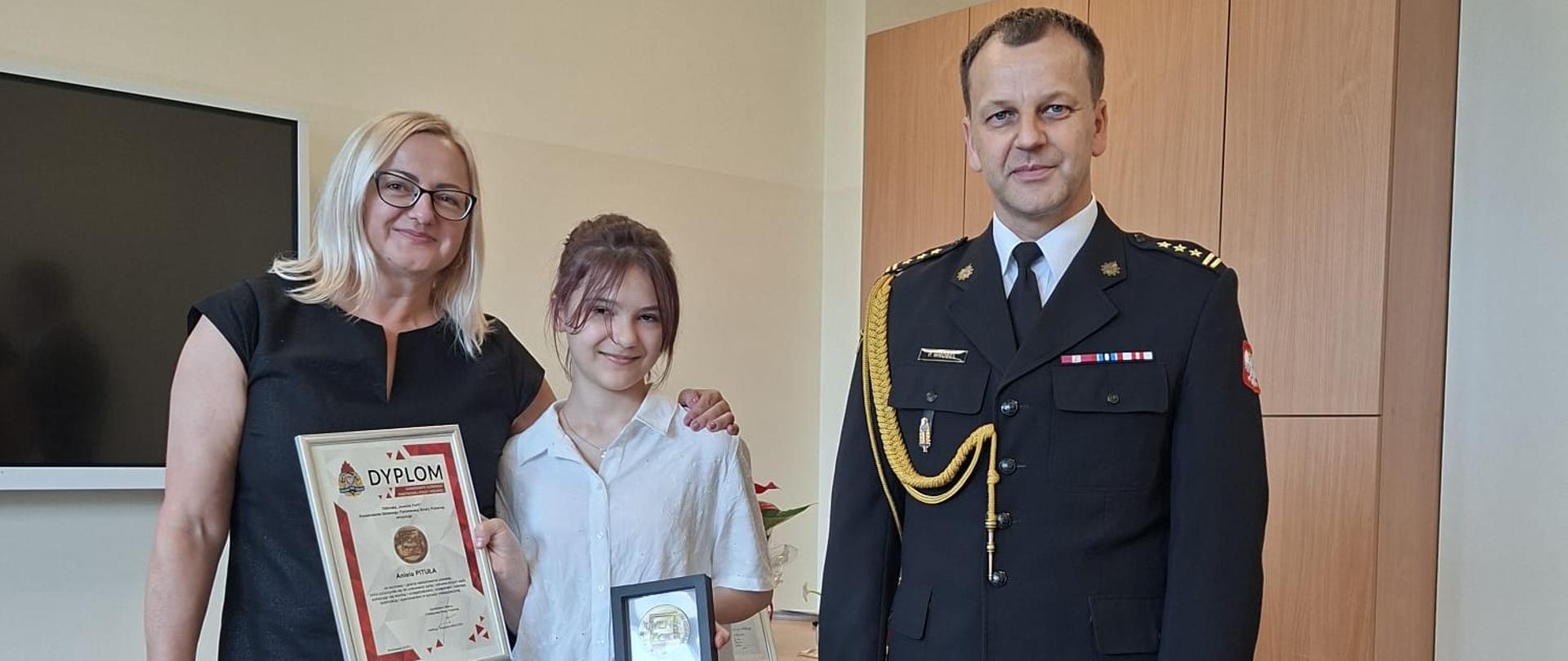 Odznaczona dziewczynka w białej bluzce stoi obok z-cy komendanta miejskiego i pani w czarnej sukience. W pomieszczeniu widać szafy oraz ekran multimedialny