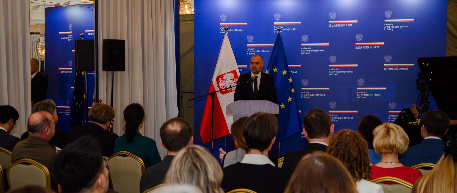 Na pierwszym planie goście, na drugim planie Ambasador Jakub Kumoch wygłaszający przemówienie