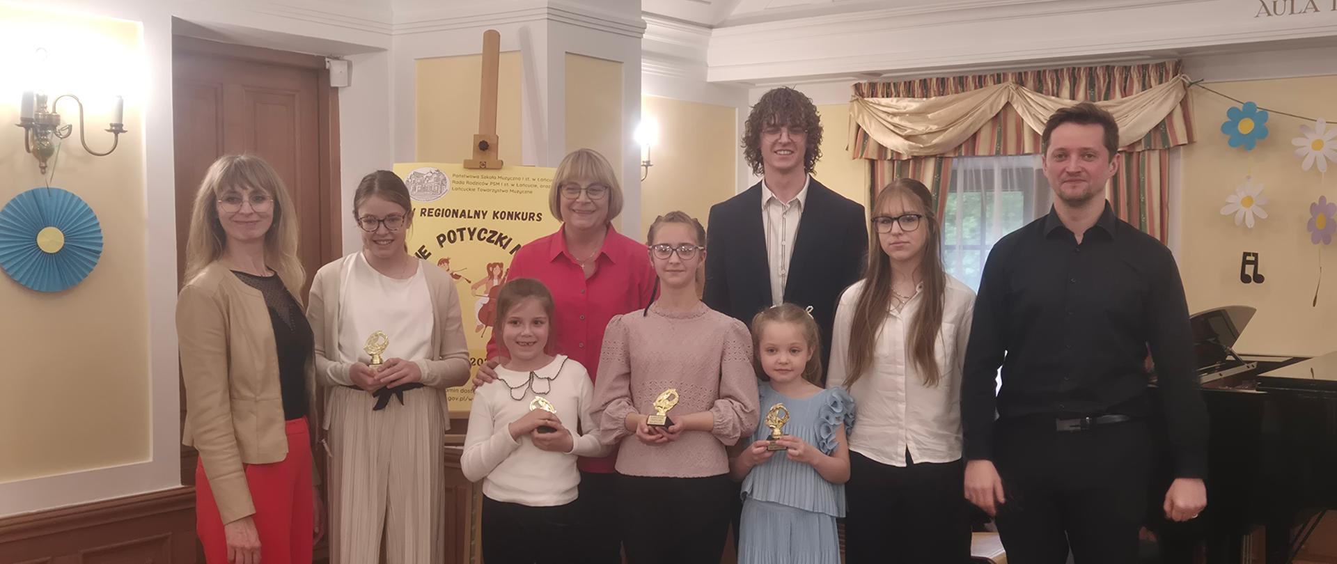 Zdjęcie grupowe wykonane w eleganckiej sali koncertowej z drewnianą boazerią i ozdobnym sufitem. Na podwyższeniu stoi dziewięć osób: czworo dorosłych oraz sześcioro dzieci (uczestników konkursu). Dzieci z dumą prezentują trzymane w dłoniach małe, złote statuetki. Na pierwszym planie, na podłodze przed grupą, leży wiolonczela wraz ze smyczkiem. W tle po lewej stronie widoczna jest niebieska dekoracja ścienna, pośrodku plakat konkursowy na sztaludze, a po prawej stronie fragment czarnego fortepianu i papierowe kwiaty na ścianie.