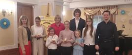Zdjęcie grupowe wykonane w eleganckiej sali koncertowej z drewnianą boazerią i ozdobnym sufitem. Na podwyższeniu stoi dziewięć osób: czworo dorosłych oraz sześcioro dzieci (uczestników konkursu). Dzieci z dumą prezentują trzymane w dłoniach małe, złote statuetki. Na pierwszym planie, na podłodze przed grupą, leży wiolonczela wraz ze smyczkiem. W tle po lewej stronie widoczna jest niebieska dekoracja ścienna, pośrodku plakat konkursowy na sztaludze, a po prawej stronie fragment czarnego fortepianu i papierowe kwiaty na ścianie.