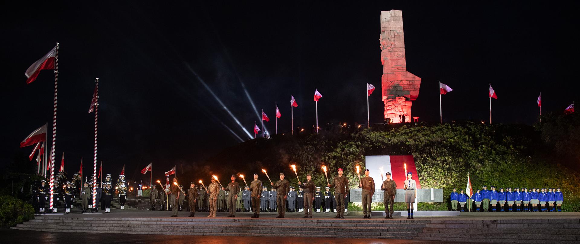 Grupa żołnierzy z pochodniami. W tle flaga biało-czerwona, podświetlony na czerwono pomnik Bohaterów Westerplatte, Wokól pomnika białe-czerwone flagi na masztach
