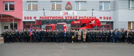 Na placu przed komendą, na tle drabiny samochodowej strażacy oraz osoby cywilne pozują do fotografii grupowej.