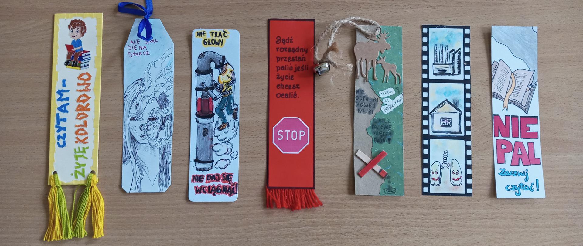 7 zakładek do książki ułożonych obok siebie na stole, każda zawiera hasło lub grafikę antytytoniową