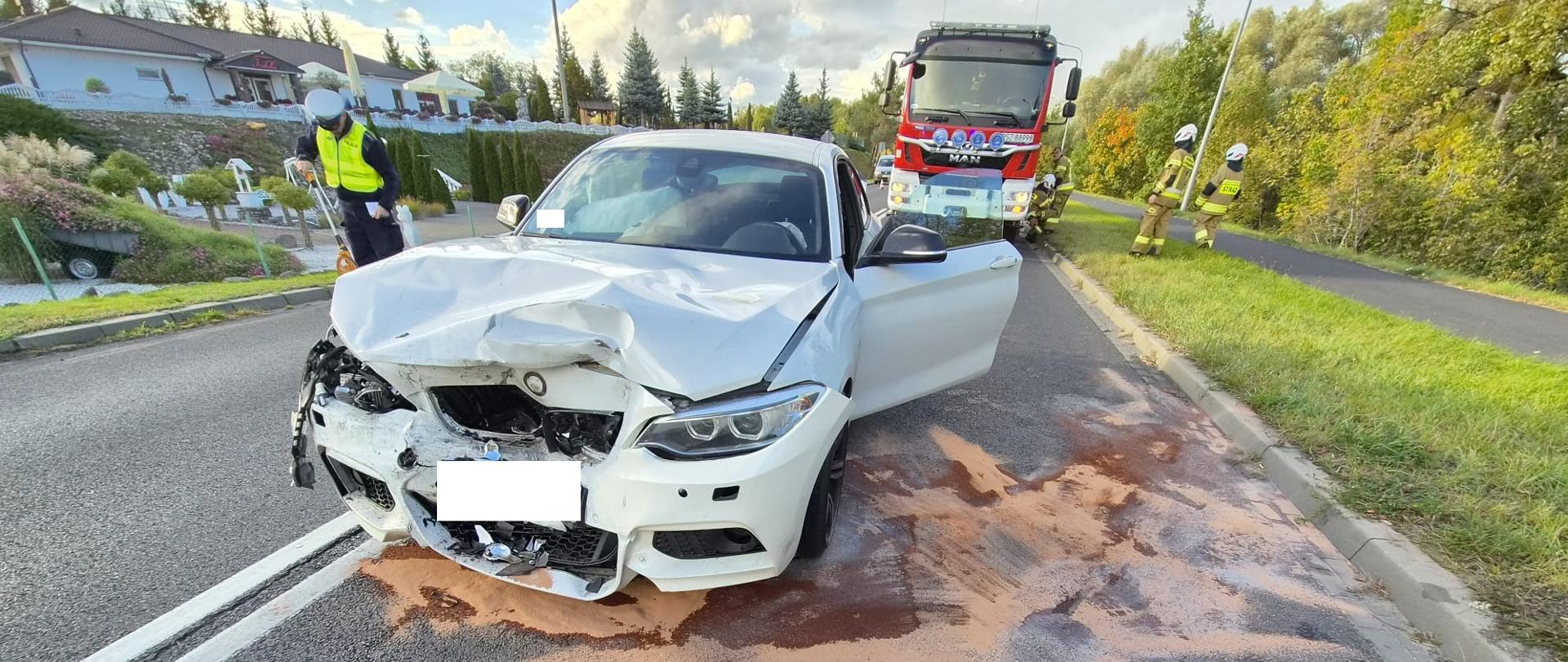 Mocno uszkodzony biały samochód na drodze, za nim samochód straży