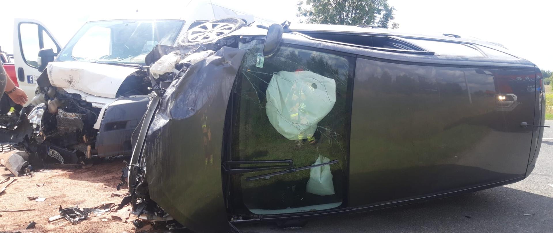 Zdjęcie przedstawia leżący na boku samochód osobowy. Za szybą widać wystrzelone poduszki powietrzne. W głębi drugi ze zniszczonych aut. 