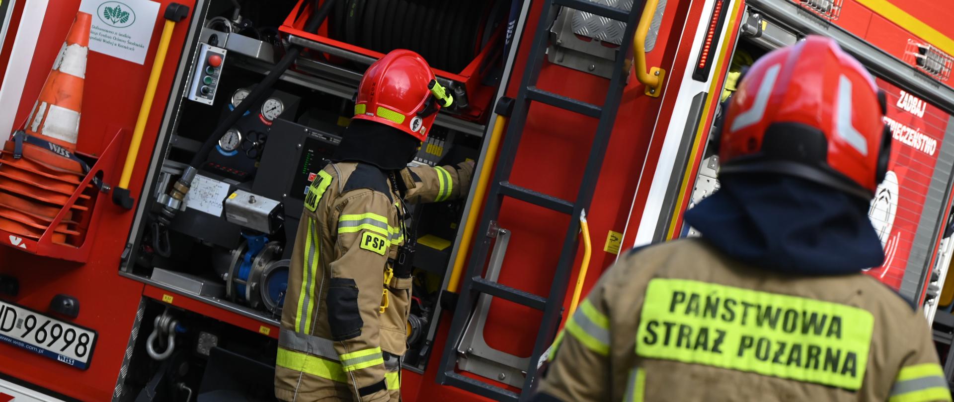 Widok z bliska na tył czerwonego wozu strażackiego marki WISS o numerze operacyjnym 663[S]21 z otwartą przestrzenią sprzętową, w której widać węże pożarnicze i panel sterowania. Po lewej stronie strażak w piaskowym mundurze z naszywką PSP i w czerwonym hełmie obsługuje aparaturę wozową. Na pierwszym planie po prawej stronie widoczne są plecy drugiego strażaka z żółtym napisem „PAŃSTWOWA STRAŻ POŻARNA”.