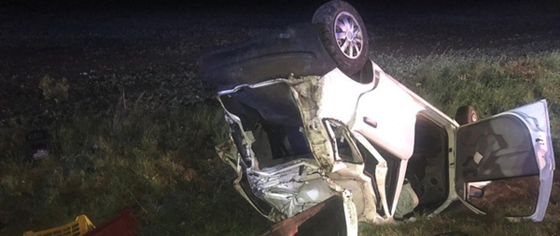 Pora nocna. Pobocze drogi oświetlone. Samochód osobowy leży na trawie na dachu z otwartymi drzwiami i tylną pogiętą klapą tylną. Za autem dwie żółte skrzynki z tworzywa sztucznego. W oddali zaorane pole uprawne.