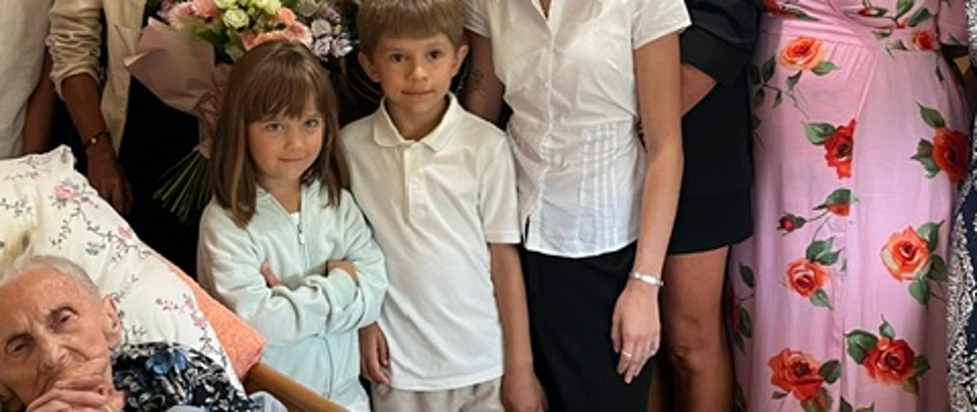 Zdjęcie przedstawia jubilatkę, panią Marię Legeżyńską, leżącą w łóżku medycznym w lewym dolnym rogu kadru i świętującą swoje 100. urodziny. Wokół stuletniej kobiety zebrali się uśmiechnięci członkowie rodziny oraz odświętnie ubrani przedstawiciele instytucji i władz lokalnych. Bezpośrednio nad łóżkiem stoją prawnuki i wnuki jubilatki, w tym młoda kobieta trzymająca duży, kolorowy bukiet kwiatów oraz pani w charakterystycznej różowej sukience w czerwone róże. 
