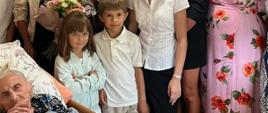 Zdjęcie przedstawia jubilatkę, panią Marię Legeżyńską, leżącą w łóżku medycznym w lewym dolnym rogu kadru i świętującą swoje 100. urodziny. Wokół stuletniej kobiety zebrali się uśmiechnięci członkowie rodziny oraz odświętnie ubrani przedstawiciele instytucji i władz lokalnych. Bezpośrednio nad łóżkiem stoją prawnuki i wnuki jubilatki, w tym młoda kobieta trzymająca duży, kolorowy bukiet kwiatów oraz pani w charakterystycznej różowej sukience w czerwone róże.