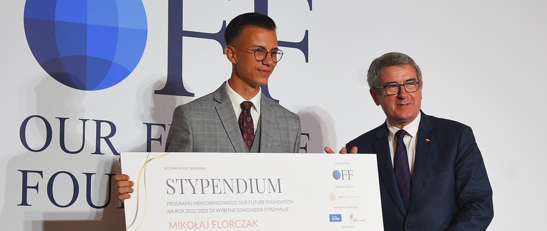 Minister Murdzek stoi obok mężczyzny w jasnym garniturze trzymającego kartkę z napisem STYPENDIUM, za nimi ścianka z napisem OFF.