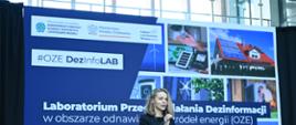 Prowadząca wydarzenie kobieta (pracownik Ministerstwa Klimatu i Środowiska) przemawia do mikrofonu na tle ekranu, na którym wyświetlany jest napis: „Laboratorium przeciwdziałania dezinformacji w obszarze odnawialnych źródeł energii (OZE)”.