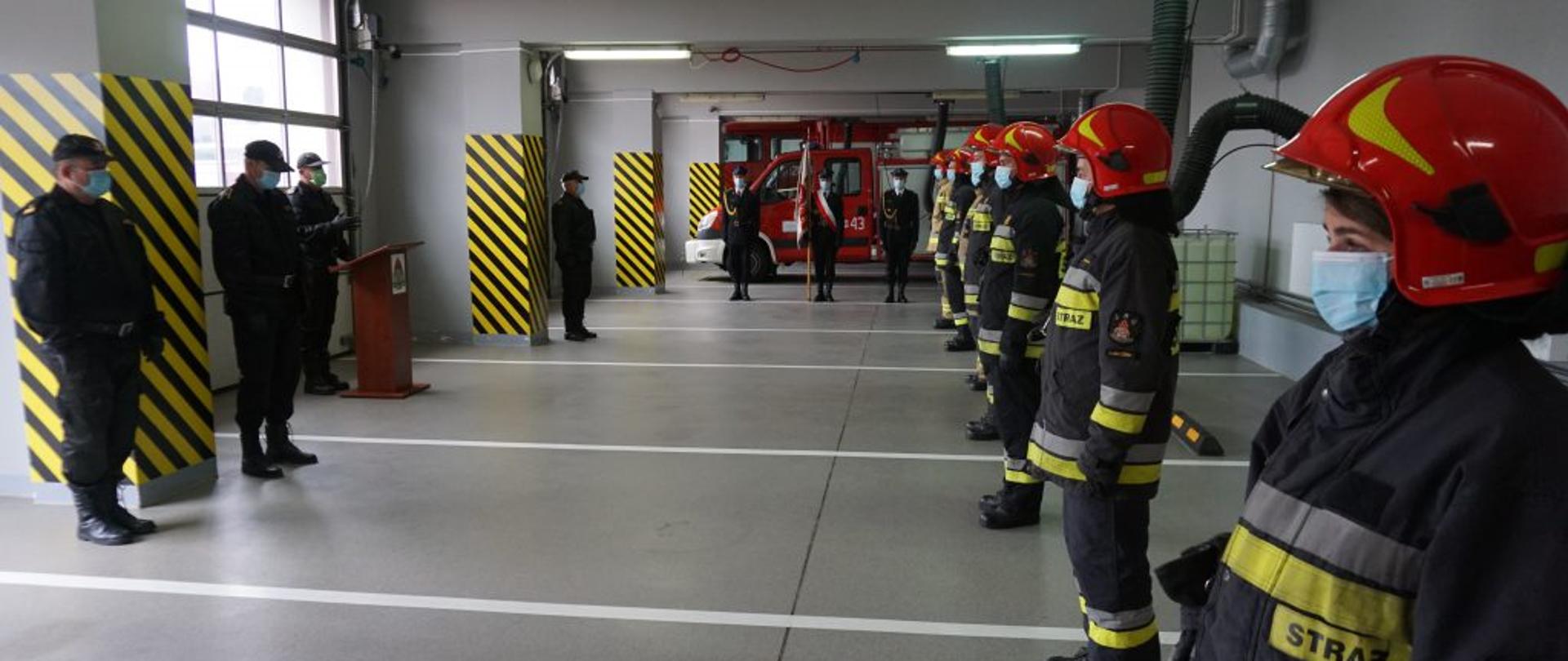 Zdjęcie zrobione w garażu Jednostki Ratowniczo-Gaśniczej w Lubaczowie. Po lewej stronie strażacy ubrani w mundury dowódczo-sztabowe. W centrum zdjęcia w oddali poczet sztandarowy, ubrany w mundur galowy. Po prawej stronie w szeregu stoją strażacy w ubraniu specjalnym. Wszyscy ubrani w maseczki ochronne. Na mównicy po lewej stronie przemawia komendant powiatowy PSP w Lubaczowie, bryg. Janusz Jabłoński.