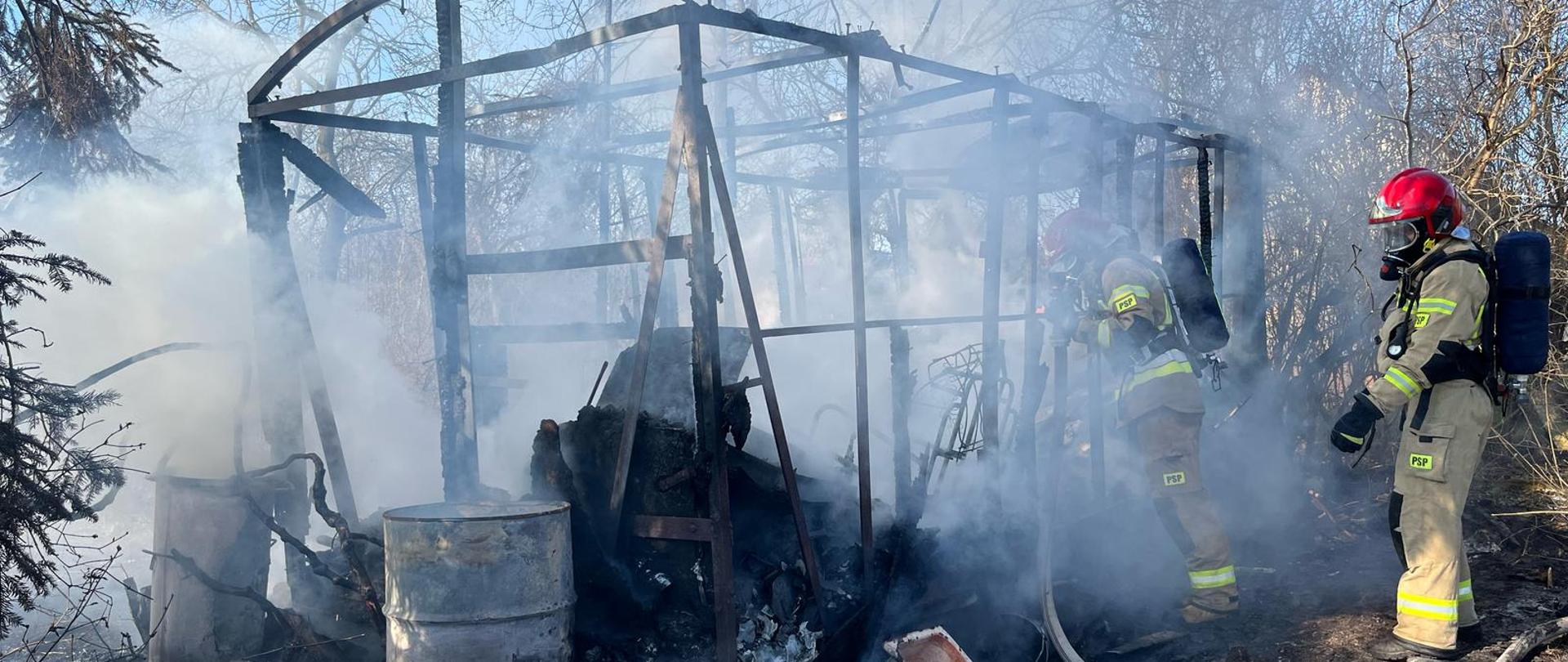 Zdjęcie przedstawia spaloną altanę działkową, z której została jedynie metalowa konstrukcja. W obrębie pogorzeliska pracuje dwóch ratowników szukających zarzewi ognia.