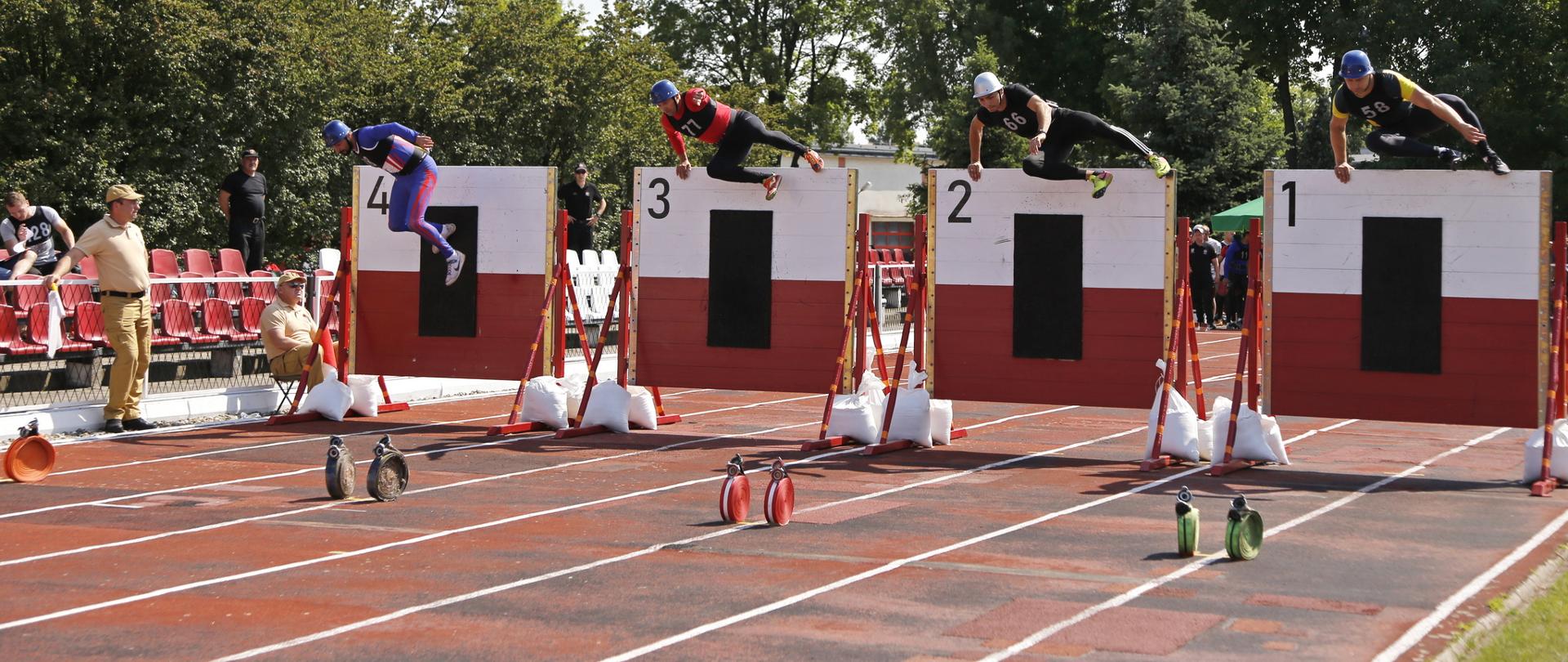 Czworo zawodników pokonujących przeszkodę jaką jest ściana, przed nimi na bieżni zwinięte węże przygotowane do zabrania po zeskoku