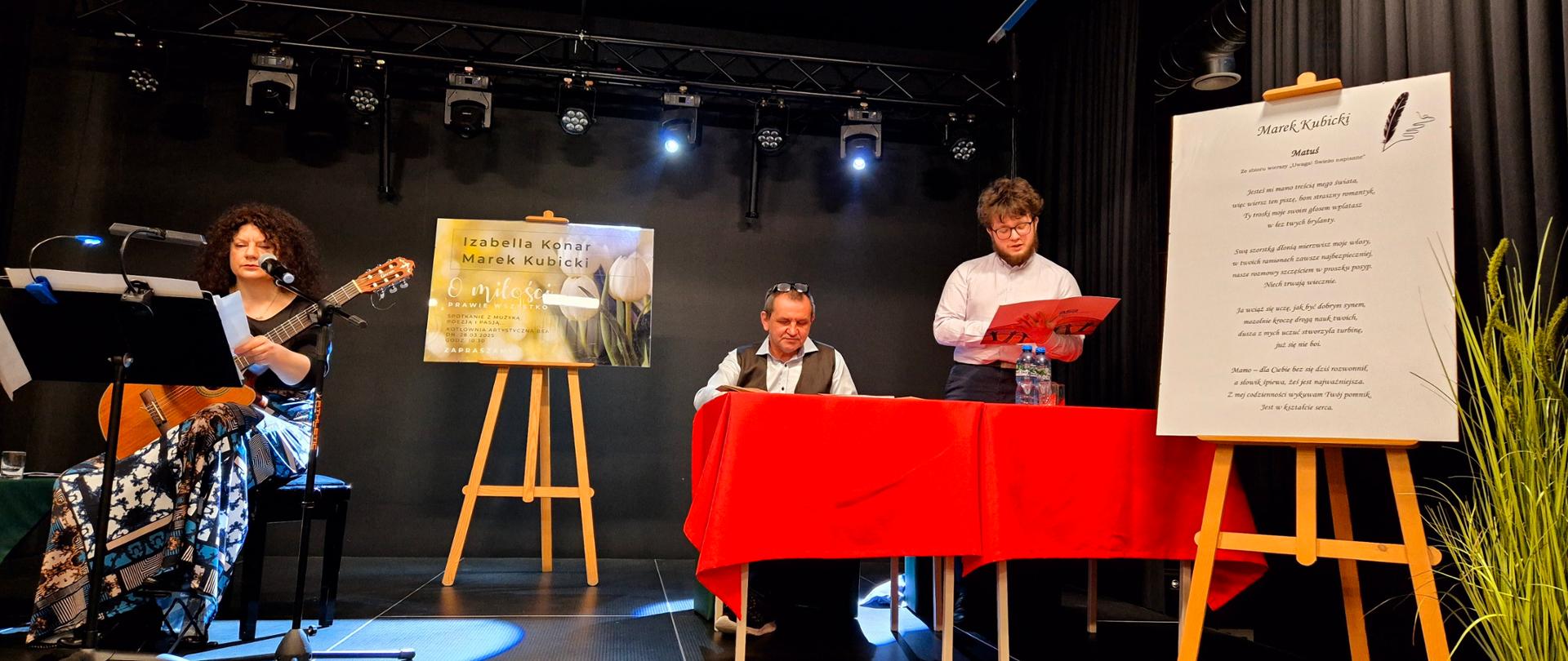 Na scenie po prawej stronie stoi roślina w doniczce, obok na sztaludze wydruk wiersza Marka Kubickiego "Matuś", dalej przy stole nakrytym czerwonym obrusem stoi mężczyzna i czyta z rozłożonej teczki, obok siedzi drugi mężczyzna. Pośrodku sceny stoi szluga z plakatem zapraszającym na koncert. Po lewej stronie siedzi kobieta z gitarą, przed nią pulpit i mikrofon.