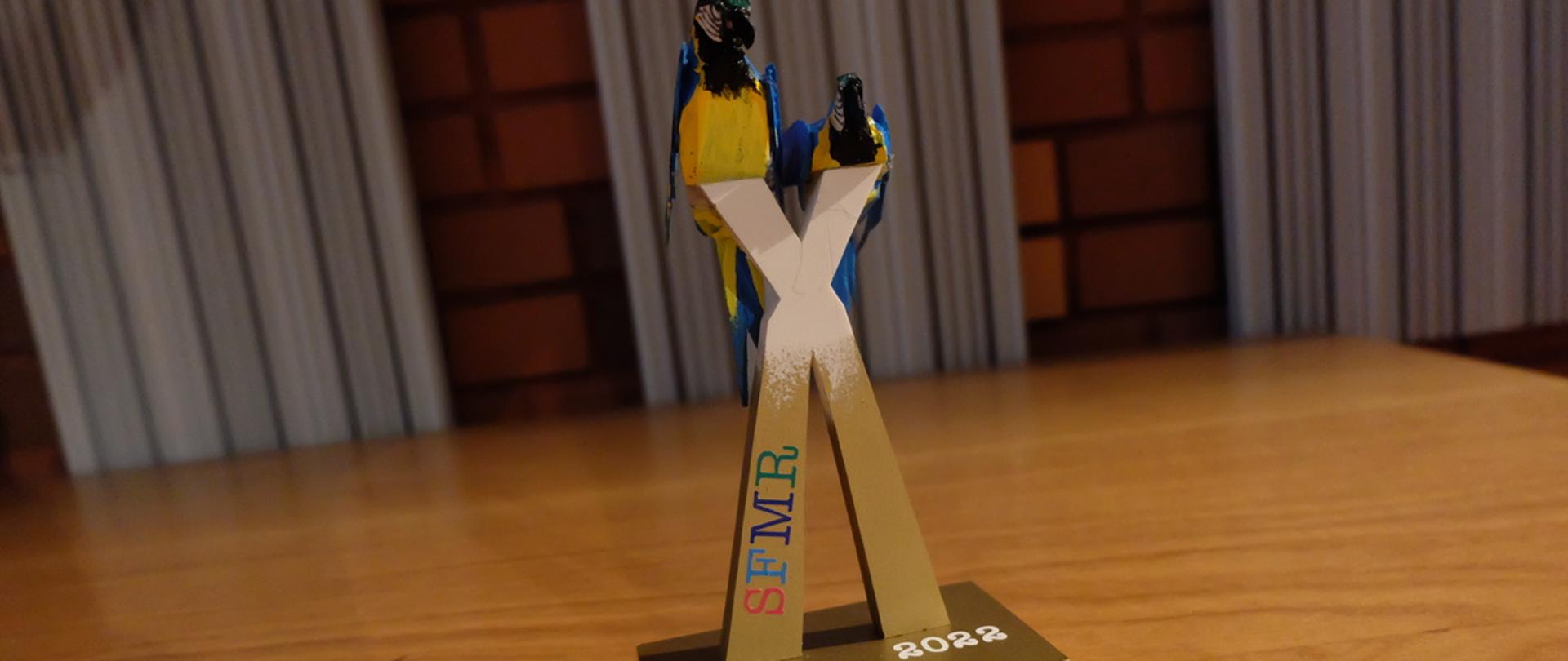 Statuetka Sieradzkiego Festiwalu Muzykujących Rodzin 2022. Na literze X siedzą 2 żółto - niebieskie papugi. Ma lewym dolnym ramieniu litery X napis SFMR, na cokole napis 2022