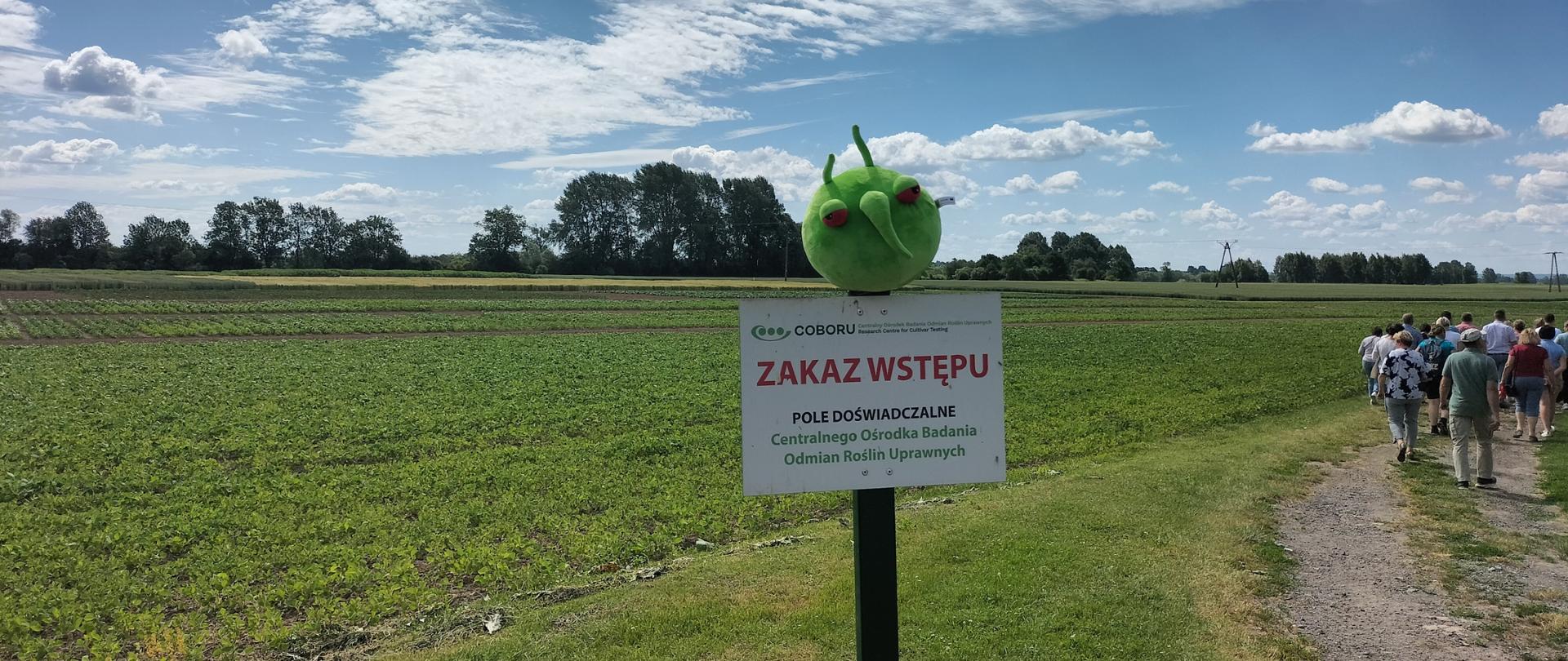 Tablica informacyjna na której siedzi zielona okrągła maskotka. W tle zielone pole, drzewa, na niebie białe chmurki. Po prawej stronie grupa ludzi idąca ścieżką.
