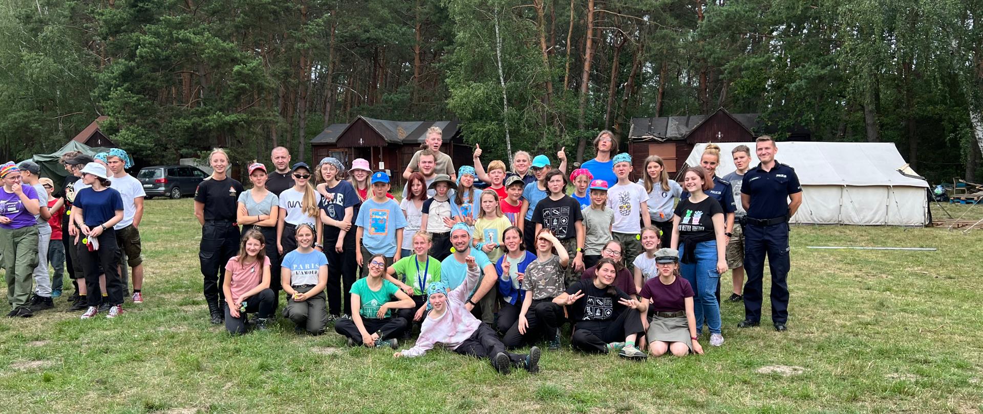 Grupa harcerzy stoi ze strażakami i policjantami na tle obozu namiotowego . Duże ciemnozielone drzewa na dalszym planie.