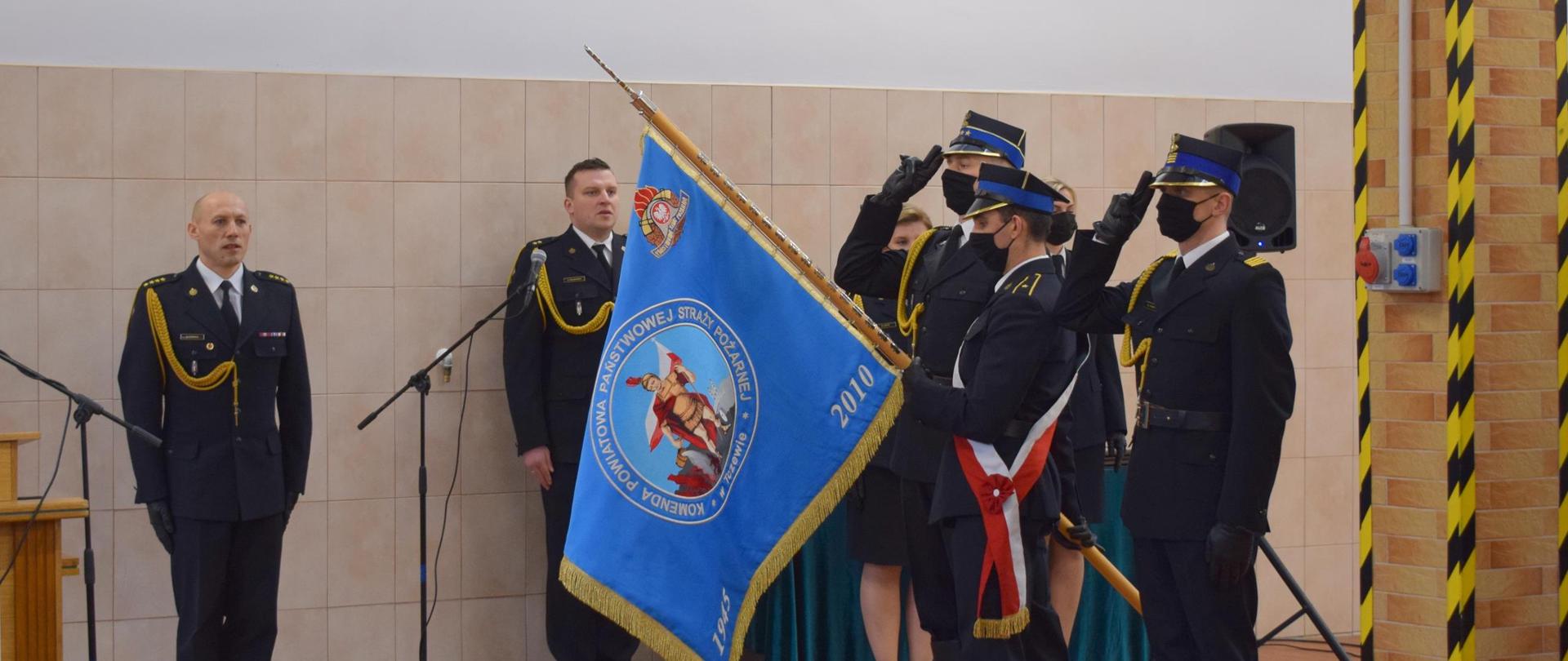 Uroczystość w garażu JRG. Moment odśpiewania hymnu. Poczet sztandarowy oddaje honor. Z tyłu dowódca uroczystości i speaker.