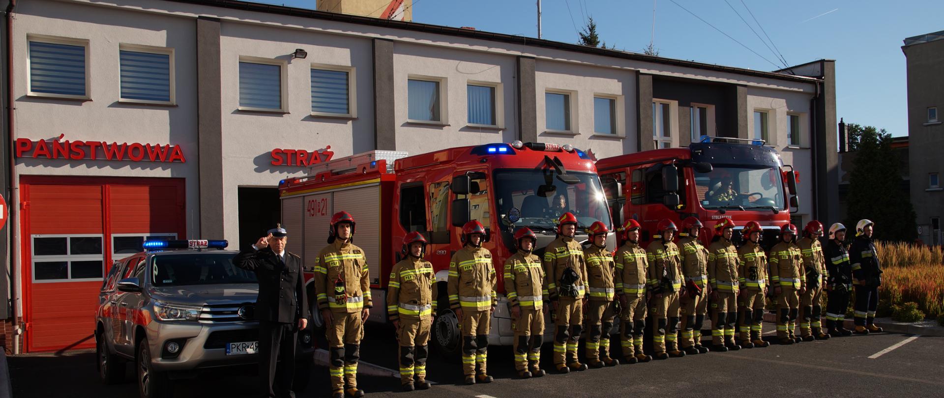 Na placu przed budynkiem komendy powiatowej stoją strażacy w ubraniach bojowych oddając hołd zmarłemu koledze, który stracił życie podczas służby. Przed budynkiem stoją samochody pożarnicze z włączonymi sygnałami dzwiękowymi i błyskowymi. 