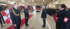 Zdjęcie przedstawia funkcjonariuszy Państwowej Straży Pożarnej w umundurowaniu wyjściowym, po stronie lewej stoi 2 funkcjonariusz, a za nimi widać płat sztandaru z napisem Bóg Honor Ojczyzna. Naprzeciw mężczyzn stoi mężczyzna w mundurze wyjściowym zwrócony twarzą w ich stronę, obok niego stoi kobieta trzymająca czerwoną teczkę w mundurze wyjściowym. 