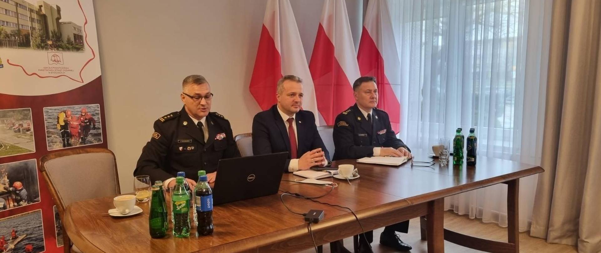 Trzy osoby siedzące przy drewnianym stole na tle trzech biało-czerwonych flag