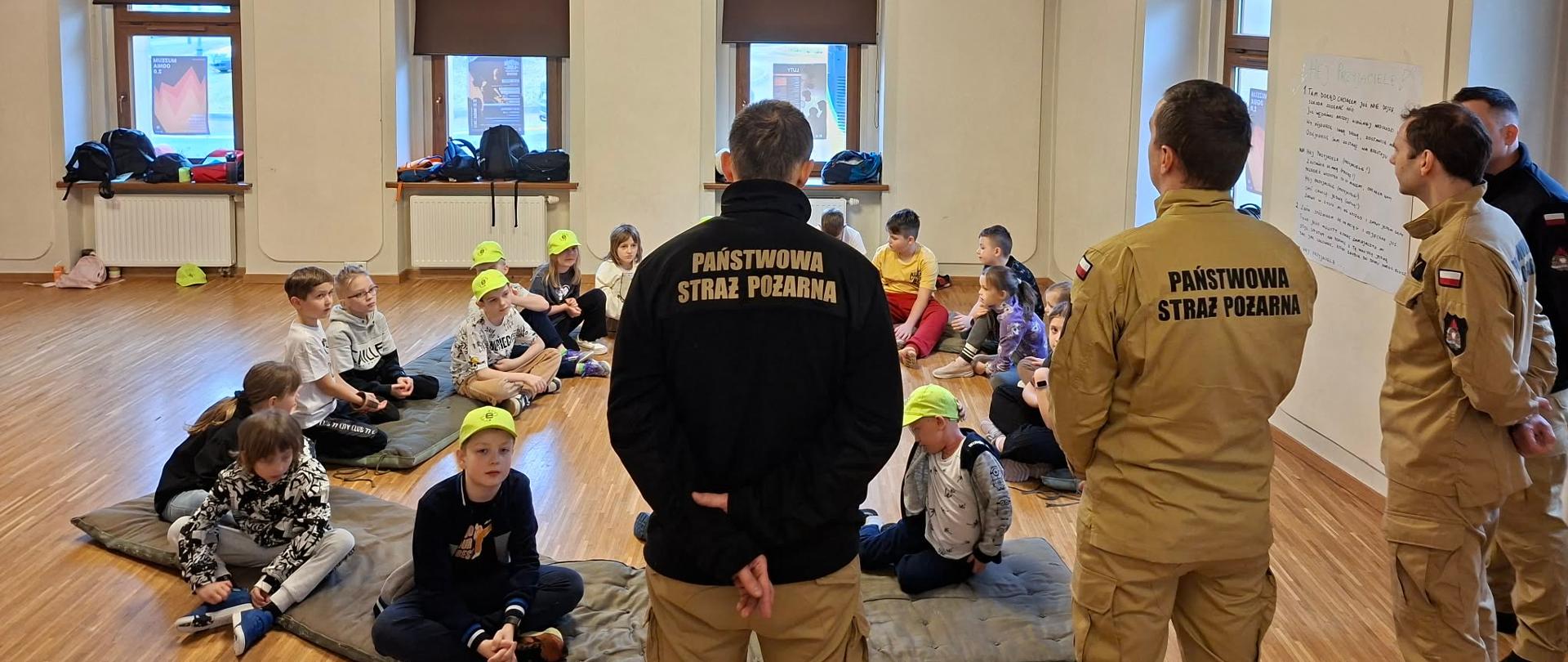 Strażacy Państwowej Straży Pożarnej podczas spotkania edukacyjnego z dziećmi. Uczestnicy półkolonii siedzą w kręgu i słuchają prelekcji dotyczącej zasad bezpieczeństwa oraz podstaw udzielania pierwszej pomocy.
