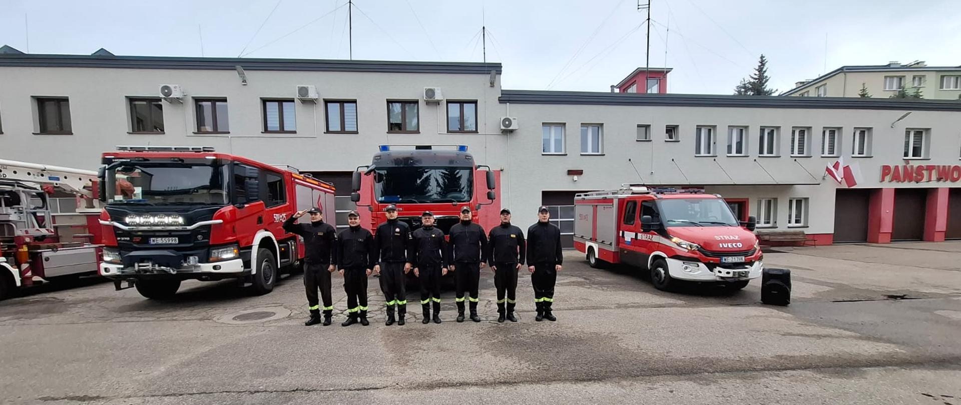 Funkcjonariusze KP PSP w Makowie Mazowieckim odśpiewują Hymn Państwowy przed budynkiem komendy. Z tyłu stoją również wozy strażackie. 