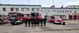 Funkcjonariusze KP PSP w Makowie Mazowieckim odśpiewują Hymn Państwowy przed budynkiem komendy. Z tyłu stoją również wozy strażackie. 
