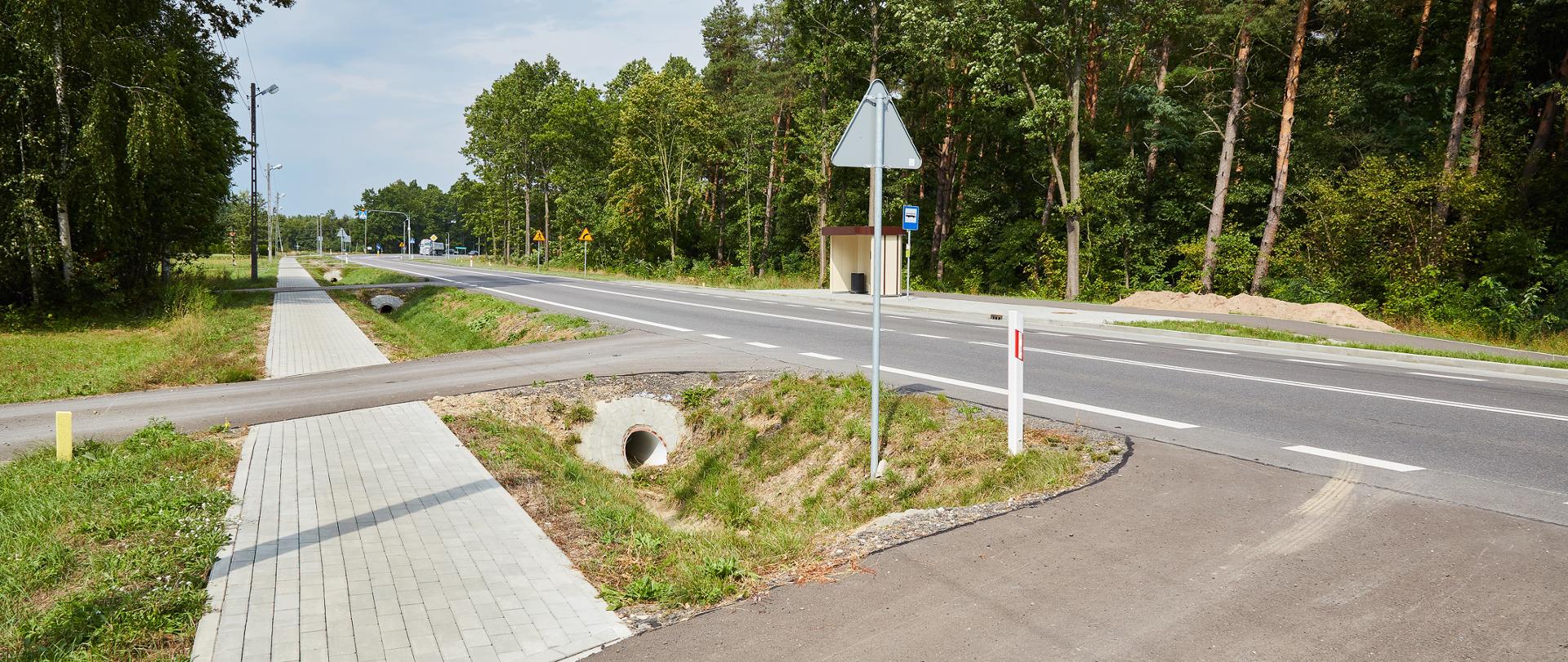 Na zdjęciu przedstawiona jest droga przebiegająca przez teren o charakterze leśnym i podmiejskim. Wzdłuż jezdni, po lewej stronie, widoczny jest nowo wykonany chodnik z kostki brukowej, prowadzący równolegle do drogi. Chodnik jest oddzielony od jezdni rowem odwadniającym, w którym znajduje się przepust z betonową rurą.
Po prawej stronie drogi znajduje się zatoka autobusowa z wiatą przystankową oraz oznakowaniem przystanku. Wzdłuż drogi ustawione są znaki ostrzegawcze i informacyjne, a także słupy oświetleniowe. Jezdnia ma wyraźne oznakowanie poziome.
Otoczenie stanowią liczne drzewa – zarówno liściaste, jak i iglaste – oraz zieleń przydrożna. W tle widoczna jest dalsza część drogi z dodatkowymi znakami i skrzyżowaniami oraz fragmentem zabudowy drogowej. Całość sprawia wrażenie uporządkowanej infrastruktury drogowej, dostosowanej do ruchu pieszych i komunikacji publicznej.