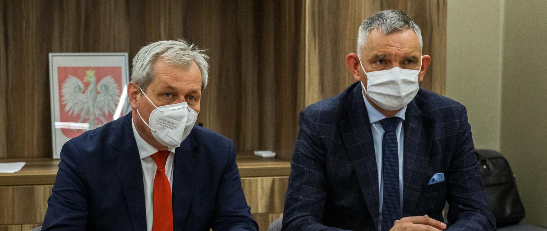 Na zdjęciu dwóch mężczyzn siedzących przy stole. Po lewej wojewoda opolski, opierający łokcie o blat stołu. W dłoniach trzyma okulary. Obok niego siedzi dyrektor USK w Opolu. Ma złożone ręce. Obaj panowie ubrani są w ciemne garnitury. Przed dyrektorem stoi procelanowa, biała filiżanka oraz szklanka. W tle widać szafę oraz zawieszone godło. 