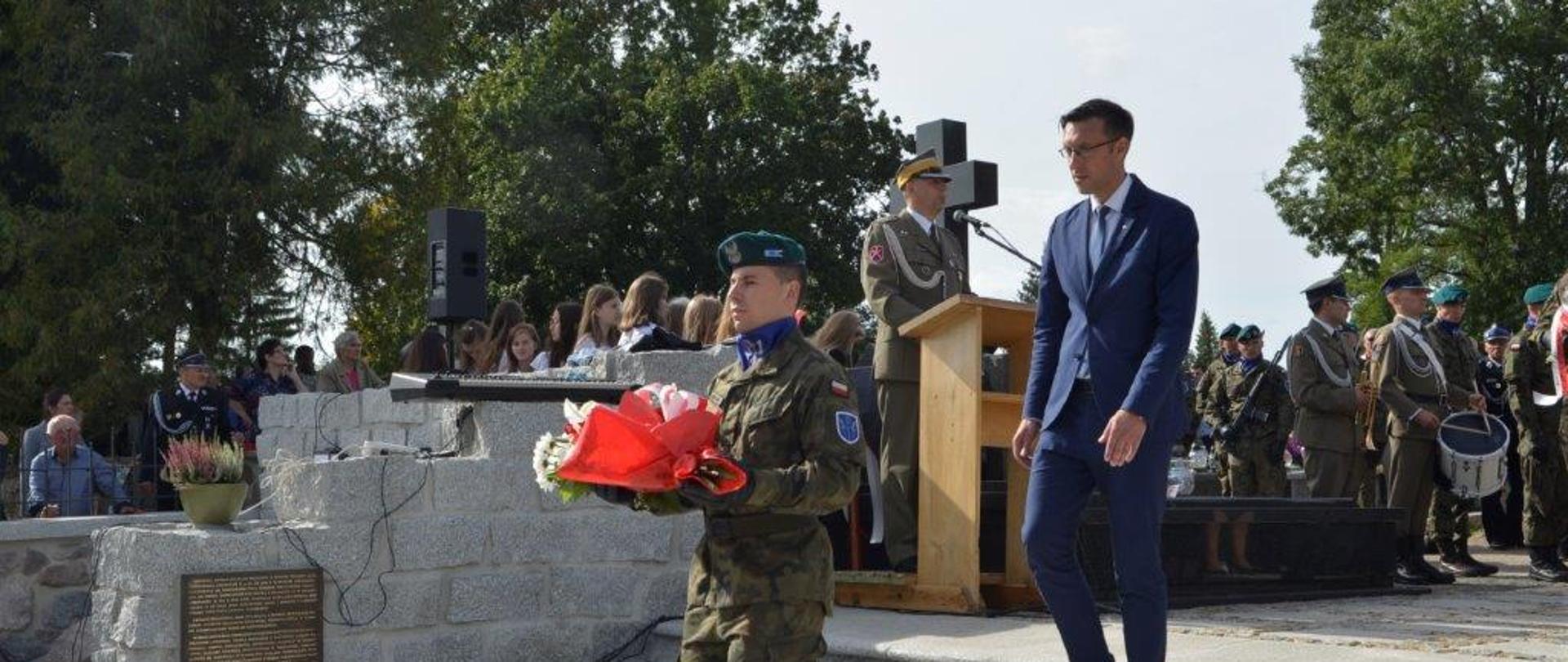 Na pierwszym planie Kierownik Delegatury Mazowieckiego Urzędu Wojewódzkiego w Ostrołęce Marcin Grabowski schodzi po kamiennych schodach. Przed nim żołnierz niesie wiązankę, która zostanie złożona w miejscu upamiętnienia poległych w czasie II wojny światowej - obiekt niewidoczny na zdjęciu. Na drugim planie kamienny murek przyklejony do schodów. Obok muru mównica, przy której przemawia żołnierz prowadzący uroczystość. W tle za mównicą i murem znajduje się grupa kilkudziesięciu osób uczestniczących w uroczystości, którzy stoją w cieniu rozłożystych drzew. 