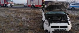 Rozbity samochód osobowy koloru białego. W tle 2 samochody pożarnicze i radiowóz policji