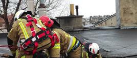 Na zdjęciu widać uszkodzony dach. Strażacy w szelkach ratowniczych i ubraniach specjalnych - kolor piaskowy przybijają papę i deski. Zachmurzenie duże. Ściana kamienicy szara. W tle kominy kamienicy.