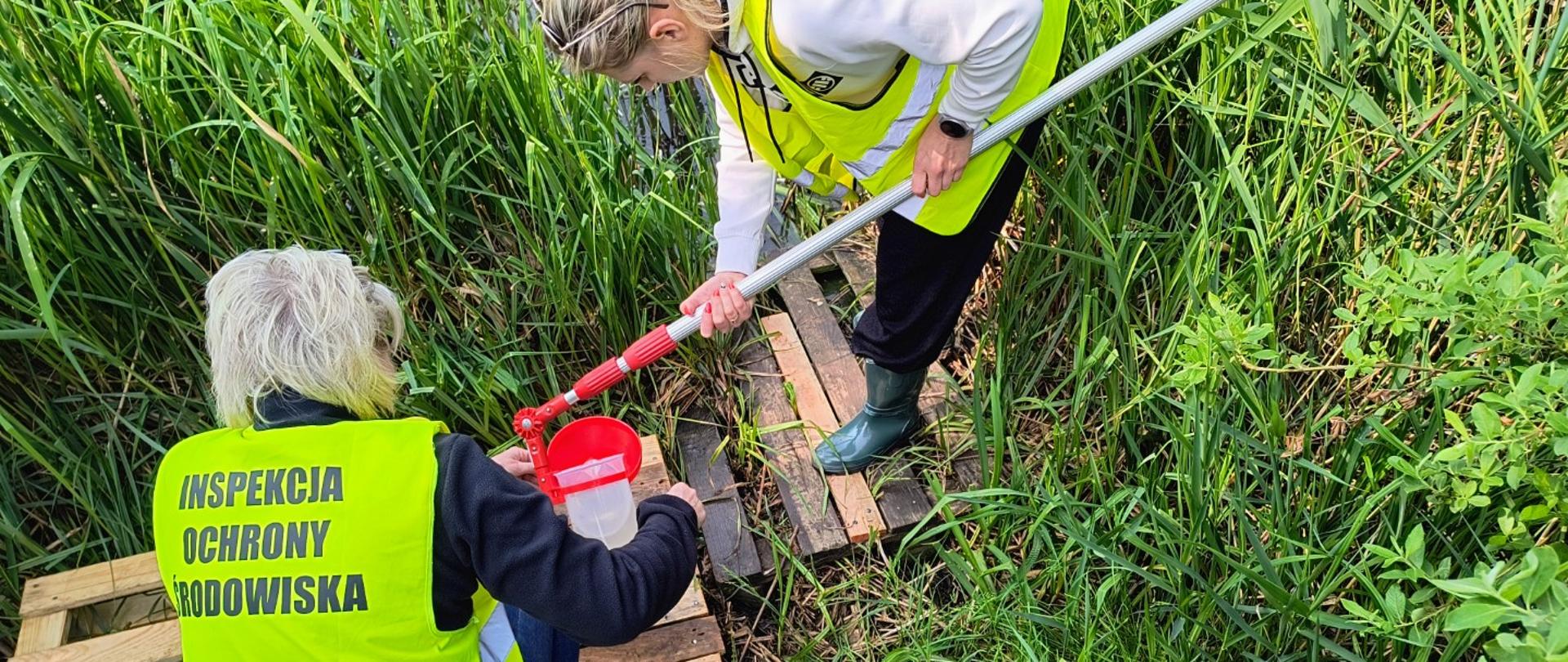 Pobór prób wód jeziora Kielno. Dwie panie w żółtych kamizelkach z napisem Inspekcja ochrony środowiska na brzegu jeziora porośniętego roślinnością. Jedna trzyma czerpak z pobraną wodą.