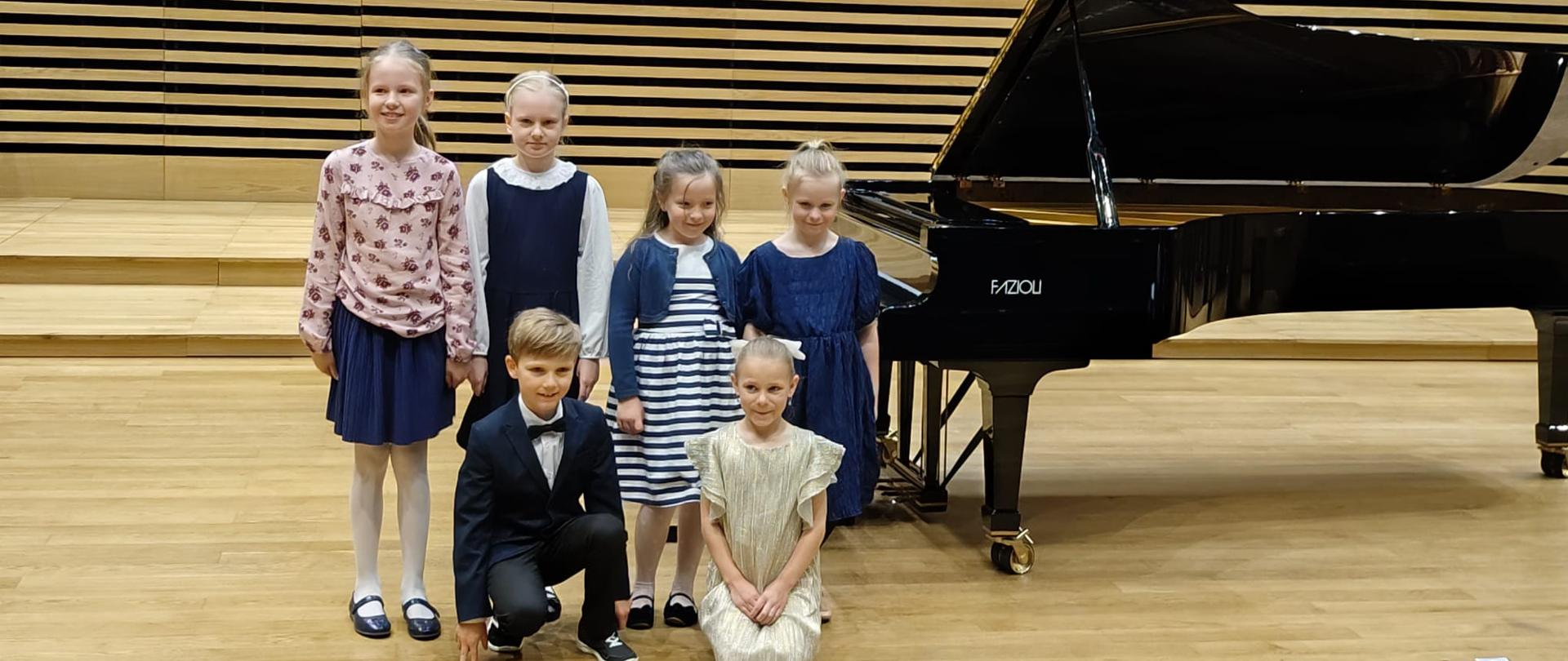 Pięć dziewczynek i chłopiec stoją na scenie przy fortepianie.