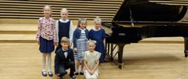 Pięć dziewczynek i chłopiec stoją na scenie przy fortepianie.