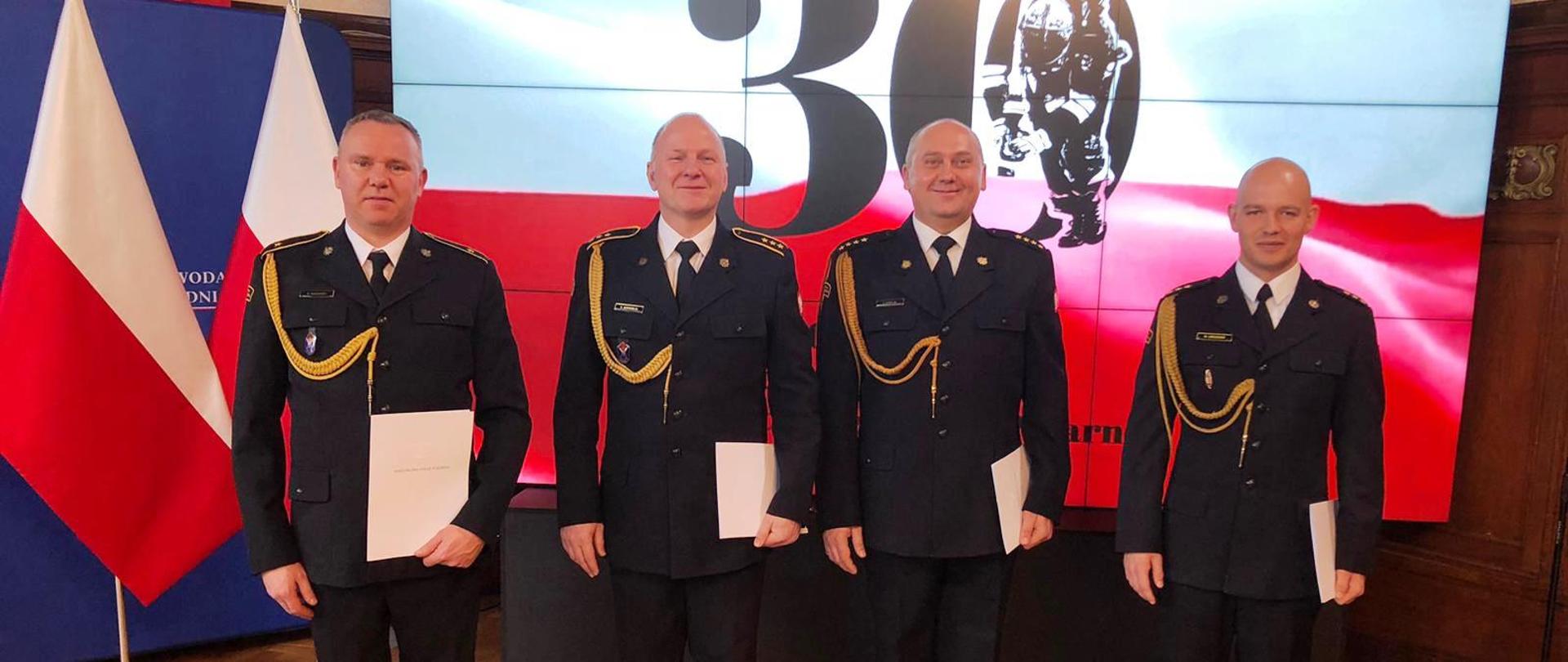 Czterech strażaków stojących w rzędzie, w ręku trzymają akty nadania awansów i nagród, w tle llogo z okazji 30-lecia PSP