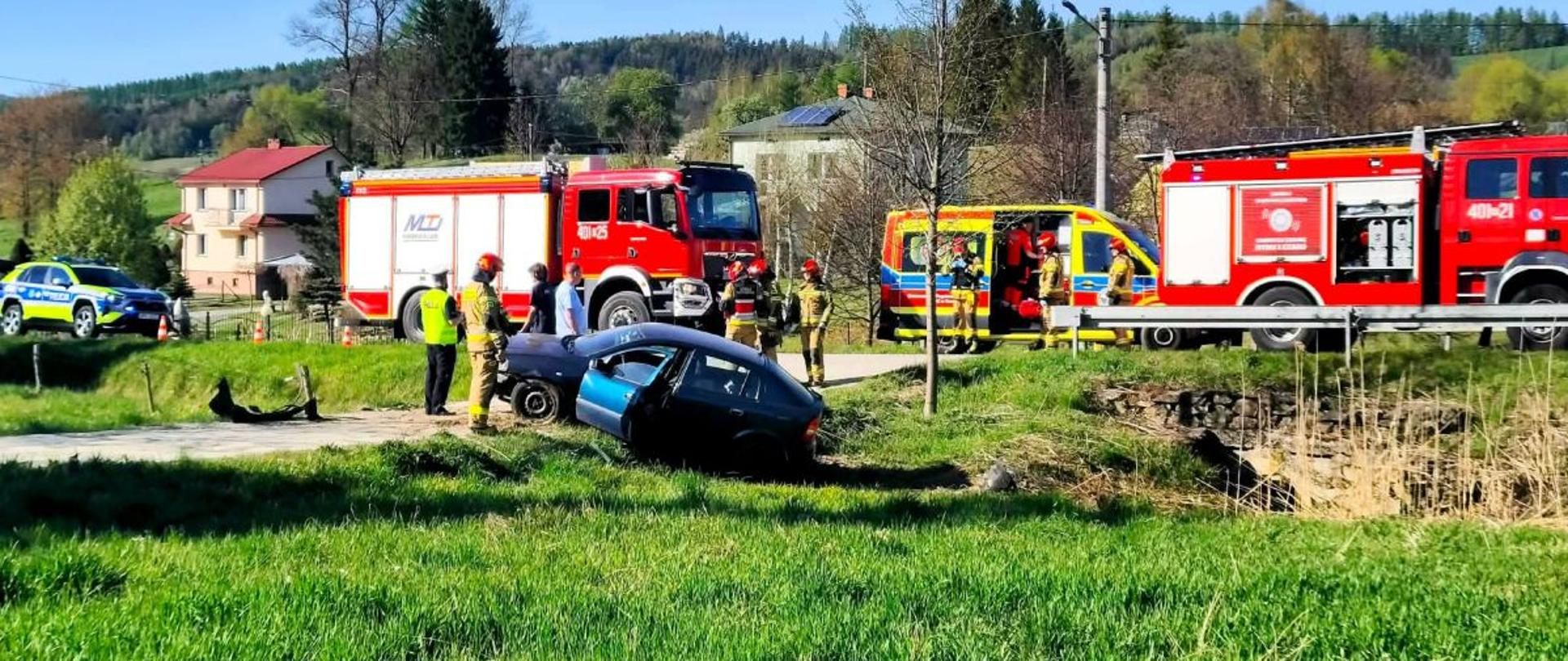 Zdjęcie przedstawia samochód osobowy poza drogą oraz dwa zastępy straży pożarnej, pogotowie i radiowóz na drodze. 