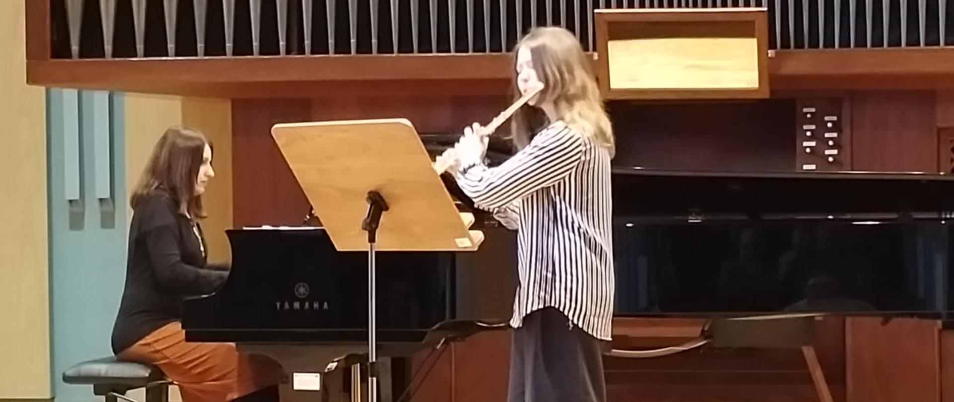 Na scenie sali koncertowej uczennica gra na flecie, przed nią pulpit. Za nią gra akompaniatorka na fortepianie. Za nimi w tle organy.