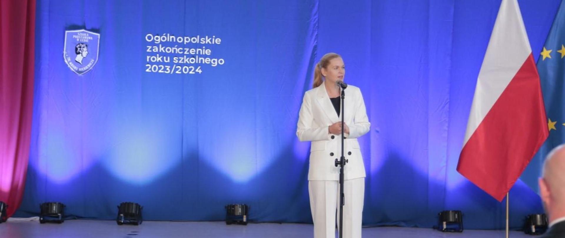 Na tle niebieskiej dekoracji z tarczą szkoły i napisem Ogólnopolskie zakończenie roku szkolnego 2024/2025 oraz flag polski i UE stoi minister Nowacka i przemawia do mikrofonu.