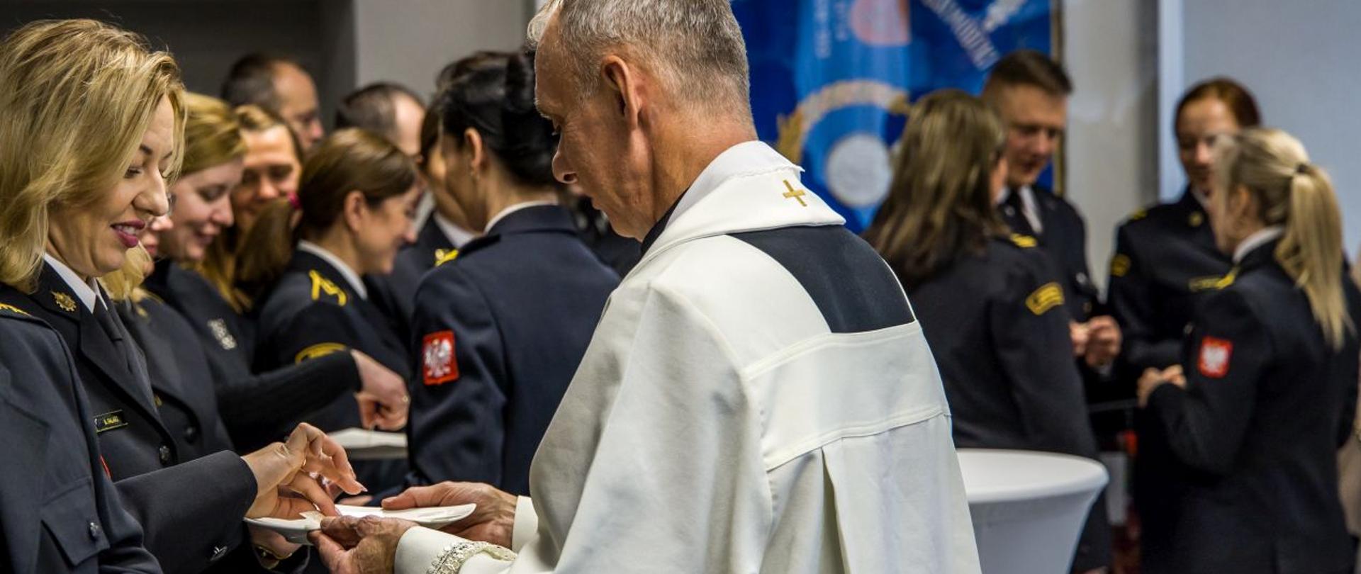 Ksiądz kapelan rozdaje opłatki dla funkcjonariuszy i pracowników cywilnych KW PSP w Opolu. W tle widoczny wiszący w gablocie sztandar KW PSP w Opolu.