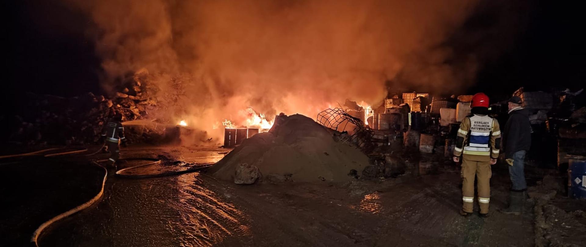 Zdjęcie przedstawia pożar składowiska odpadów, strażacy podają jeden prąd gaśniczy wody w natarciu na palące się składowisko. Po drugiej stronie stoi strażak w kamizelce z napisem Kierujący Działaniem Ratowniczym z osobą postronną.