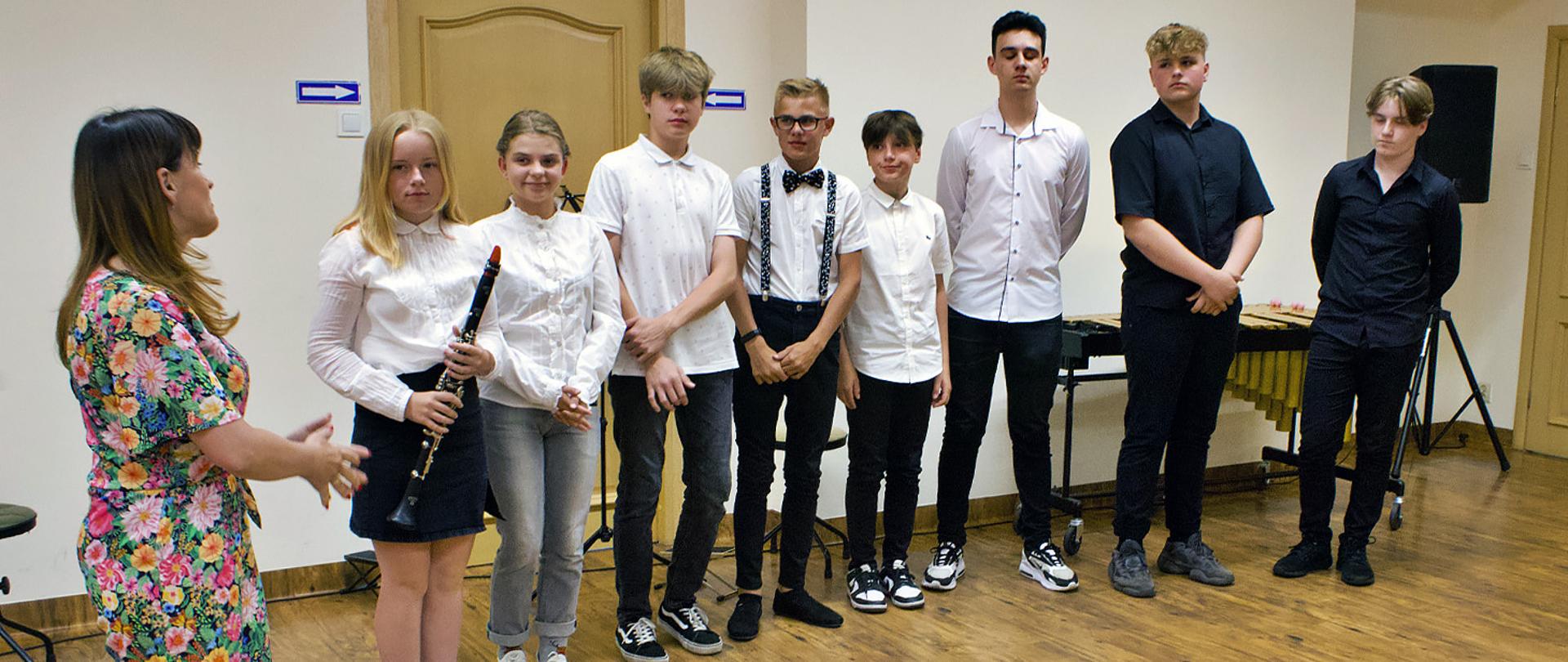 Na zdjęciu grupa osób stojąca w auli szkoły. Z lewej strony nauczycielka stojąca bokiem i patrząca w prawą stronę na grupę 8 uczniów - 2 dziewczynki i 6 chłopców. Jedna z dziewczynek trzyma w ręku klarnet, za uczniami stoi ksylofon.