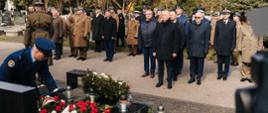 Uczestnicy ceremonii upamiętniającej składają wieńce i oddają hołd przy grobie na cmentarzu wojskowym; wśród obecnych znajdują się przedstawiciele służb mundurowych w galowych mundurach, w tym Wojska Polskiego, Straży Pożarnej, Policji i innych formacji.