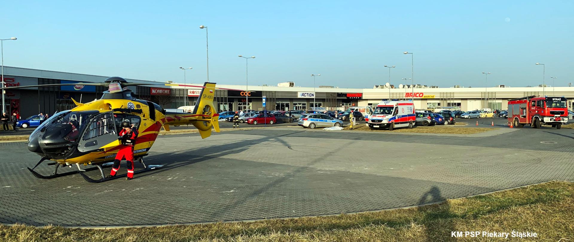 Zdjęcie przedstawia od lewej śmigłowiec LPR, radiowóz policji, karetkę ZRM oraz średni samochód ratowniczo - gaśniczy. Z tyłu widoczne jest centrum handlowe.