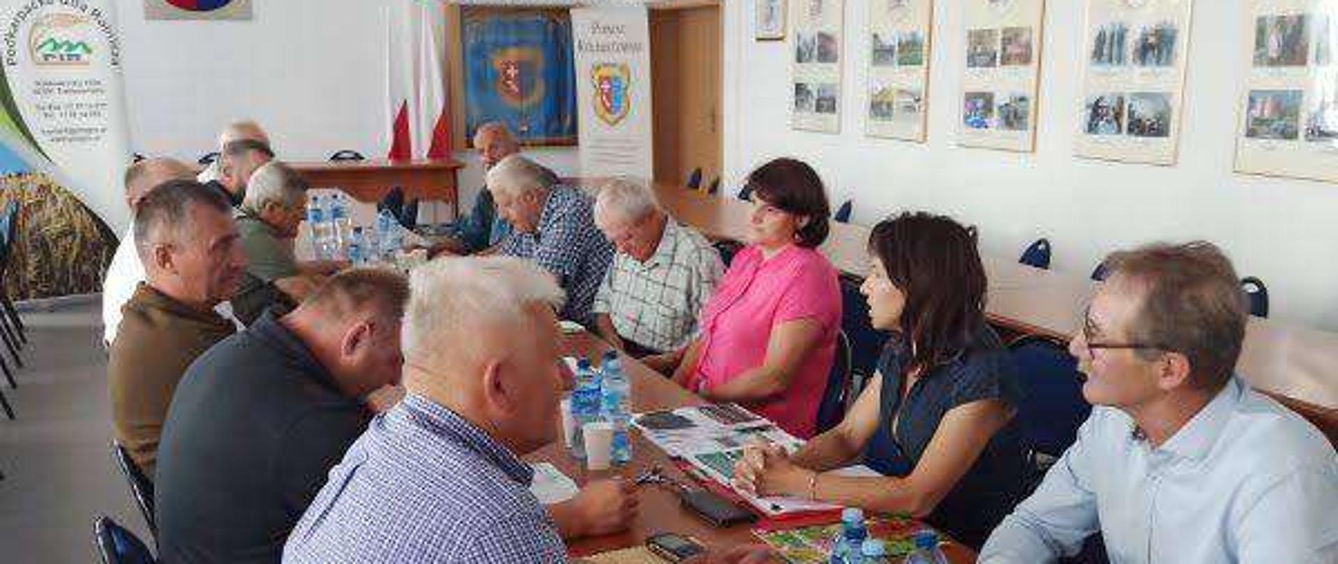 Grupa osób przy długim stole konferencyjnym. Siedzą naprzeciwko siebie i rozmawiają.