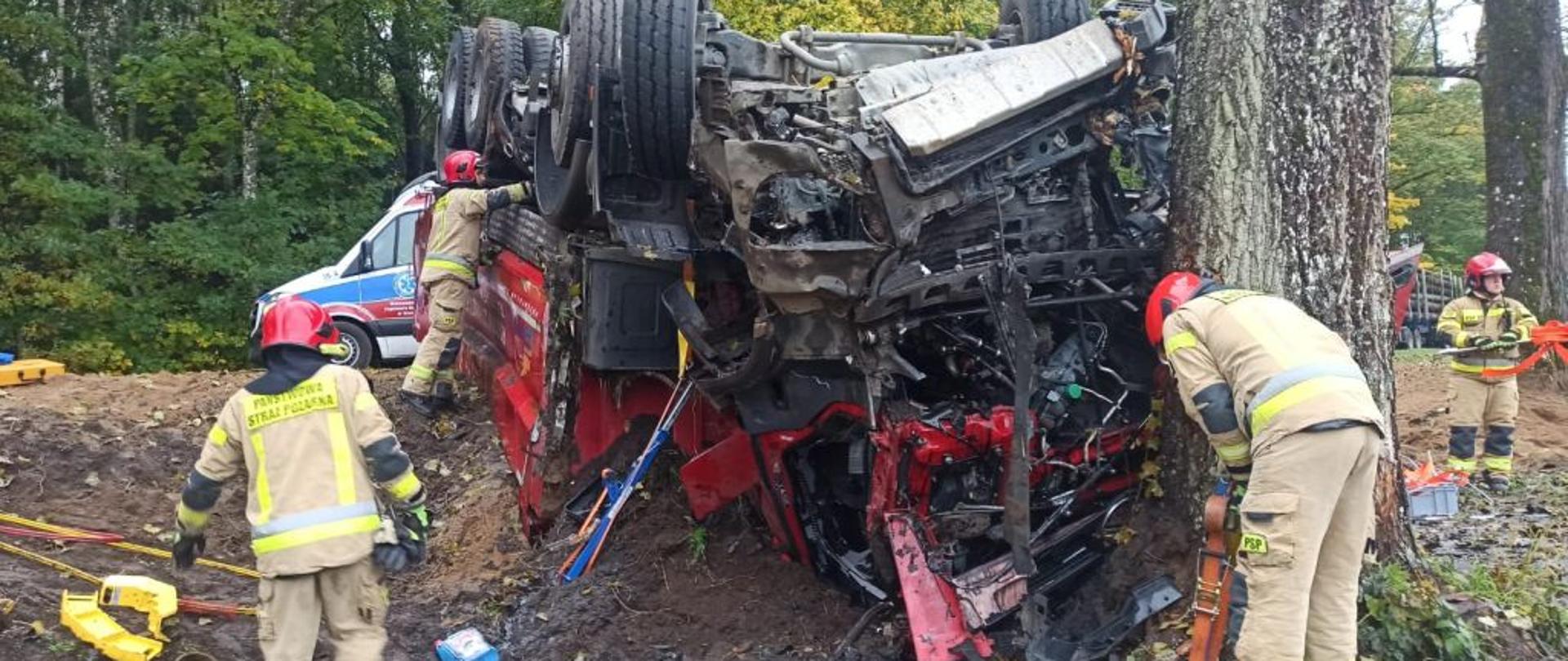 Zbliżenie na leżącą ciężarówką. Ciężarówka leży na dachu, ma zniszczony przód, zgniecioną kabinę. Przy aucie stoją strażacy.